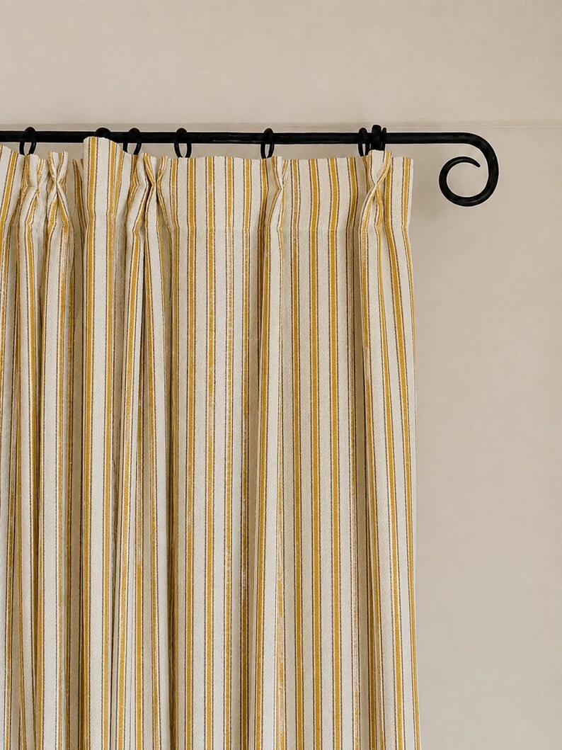 May include: A pair of off-white curtains featuring vertical mustard yellow stripes, suspended from a black metal rod with decorative scroll ends. The fabric is gathered into pleats at the top, creating a series of uniform vertical folds.