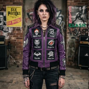 Puede incluir: Una chaqueta de cuero morada con tachuelas metálicas plateadas en hombros y cuello. El frente muestra seis parches cuadrados etiquetados como Flaming Skull, Serpent, Tribal Wolf, Dragon Eye, Moon Bat y Gothic Rose, junto a parches de calaveras.
