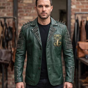 May include: A dark green leather jacket featuring silver-toned metal pyramid studs along the shoulders, lapels, and sleeves. The jacket includes a gold-toned embroidered cobra patch on the left chest, a three-button front closure, and zippered cuffs.