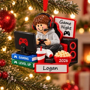 May include: A Christmas tree ornament featuring a figure wearing a gray hoodie and red headphones, holding a game controller. The figure sits before a computer monitor, stacked books labeled GAMING and LEVEL UP, a drink, popcorn, and a sign reading Logan 2026.