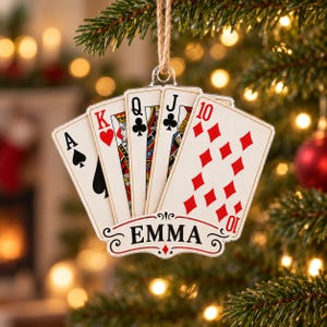 May include: A flat, fan-shaped Christmas tree ornament featuring a royal flush of playing cards including the Ace of Spades, King of Hearts, Queen of Clubs, Jack of Clubs, and 10 of Diamonds. A white banner at the bottom displays the name EMMA.