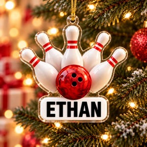 May include: A clear acrylic Christmas ornament featuring a group of white bowling pins with red stripes and a red bowling ball in the center. Below the bowling pins is a white rectangular plaque with the name ETHAN printed in bold black letters.