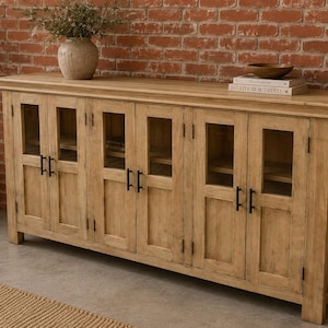 Puede incluir: Un aparador largo de madera de tono claro con seis puertas de armario que tienen paneles de cristal y tiradores de metal negro. La unidad cuenta con una superficie superior rectangular y una base sólida, ofreciendo espacio de almacenamiento tras las puertas acristaladas.