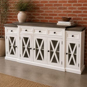 May include: A long white sideboard with a dark wood top, featuring five drawers with small black knobs and five cabinet doors with glass panels and white X-shaped mullions. The unit has a distressed finish and sits on a flat base.