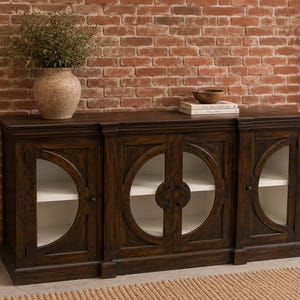 May include: A dark brown wooden sideboard featuring three cabinet sections with circular glass panels and white interior shelving. The central cabinet has double doors with circular wooden accents, while the two side cabinets have single doors.