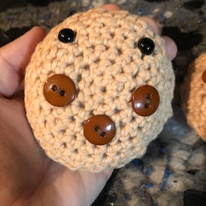 May include: A crocheted beige stuffed animal with brown button eyes and brown buttons for a nose and mouth.