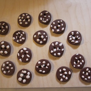 May include: A collection of 14 brown ceramic buttons with white polka dots. The buttons are arranged in a semi-circle on a light brown wooden surface.