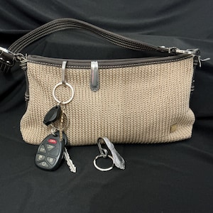 May include: A beige woven handbag with dark brown leather trim and a single shoulder strap. A silver metal spoon handle is attached to the top edge. A key ring with a black car key fob and a separate silver spoon handle key ring are included.