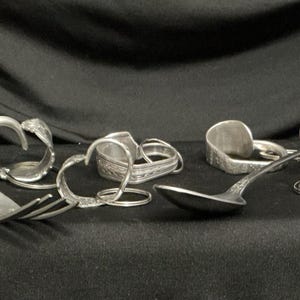 Könnte beinhalten: Eine Sammlung von silberfarbenen Metallgegenständen aus wiederverwertetem Besteck, darunter eine Gabel, ein Löffel und mehrere runde Schlüsselanhänger mit gebogenen herzförmigen Designs und geprägten Mustern auf den Griffen.