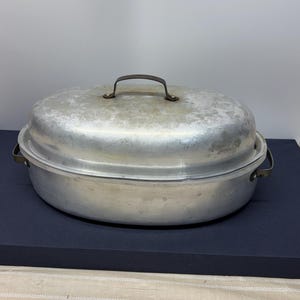 Könnte beinhalten: Ein ovaler Bräter aus Aluminium mit passendem Deckel. Der Bräter verfügt über zwei seitliche Griffe und einen einzelnen gewölbten Griff auf dem Deckel, die alle aus dunklem Metall gefertigt sind. Die Außenfläche weist Gebrauchsspuren auf.