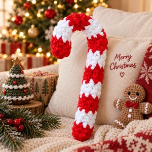 May include: A large, handmade crochet candy cane featuring alternating thick stripes of bright red and white yarn. The textured, soft-looking candy cane is positioned upright against a beige throw pillow embroidered with the text Merry Christmas.