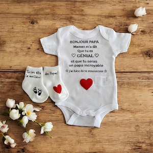 May include: A white baby bodysuit with short sleeves and black text that reads BONJOUR PAPA Maman m'a dit Que tu es GÉNIAL et que tu seras un papa incroyable j'ai hâte de te rencontrer, featuring a red heart, paired with white baby socks.
