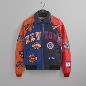 Puede incluir: Una chaqueta universitaria de cuero multicolor con paneles naranjas, azules y azul marino. El frente muestra parches bordados que incluyen el texto NEW YORK, KITH y varios logotipos de baloncesto. Tiene un cuello camisero y pu&ntilde;os acanalados.