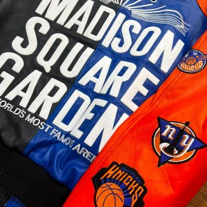 Puede incluir: Una chaqueta universitaria de cuero multicolor con letras blancas grandes que dicen MADISON SQUARE GARDEN WORLD'S MOST FAMOUS ARENA. El panel trasero es negro y azul, mientras que la manga es naranja con logotipos bordados de los New York Knicks.