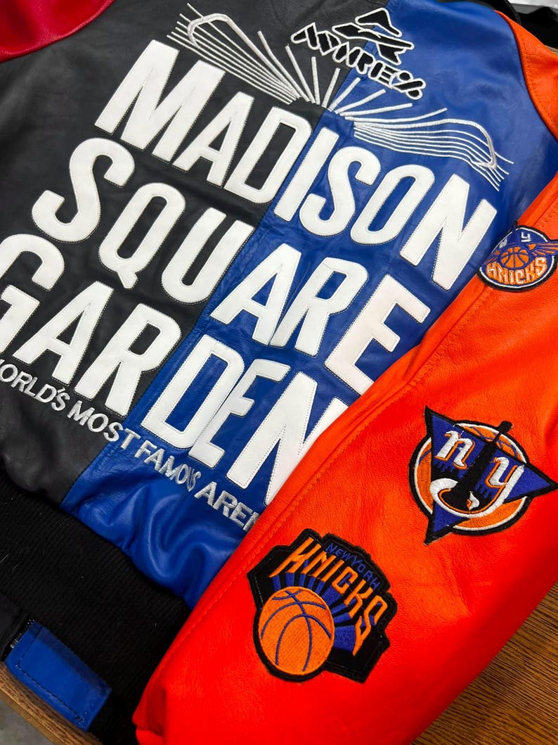 Puede incluir: Una chaqueta universitaria de cuero multicolor con letras blancas grandes que dicen MADISON SQUARE GARDEN WORLD'S MOST FAMOUS ARENA. El panel trasero es negro y azul, mientras que la manga es naranja con logotipos bordados de los New York Knicks.