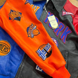 Puede incluir: Una chaqueta de cuero multicolor con paneles en naranja, azul, negro y rojo. La chaqueta muestra varios parches del equipo de baloncesto New York Knicks, incluyendo gr&aacute;ficos de balones y texto. Tiene una etiqueta blanca con la palabra LEATHER.