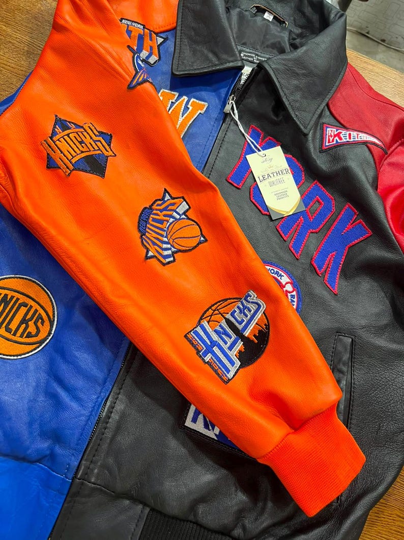 Puede incluir: Una chaqueta de cuero multicolor con paneles en naranja, azul, negro y rojo. La chaqueta muestra varios parches del equipo de baloncesto New York Knicks, incluyendo gr&aacute;ficos de balones y texto. Tiene una etiqueta blanca con la palabra LEATHER.