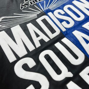 Puede incluir: Una chaqueta de cuero negra y azul con grandes letras blancas bordadas que dicen MADISON SQUARE GARDEN. Sobre el texto hay un logotipo blanco bordado con l&iacute;neas radiantes. La chaqueta tiene una textura suave y un contraste llamativo.