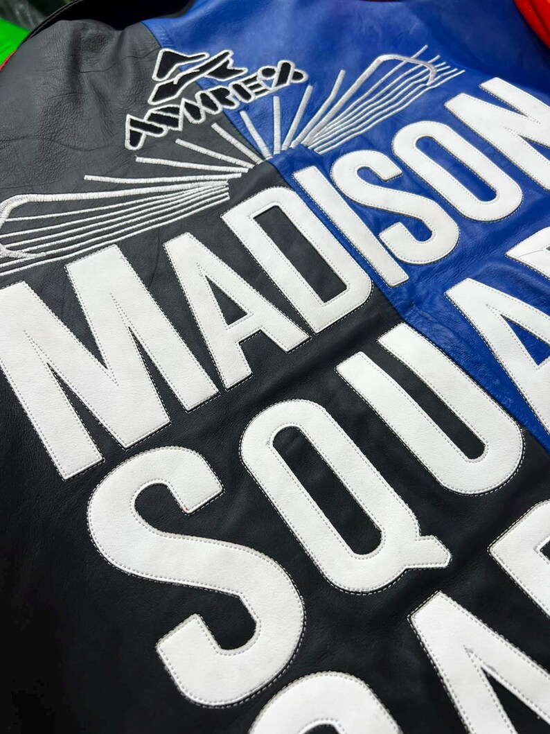 Puede incluir: Una chaqueta de cuero negra y azul con grandes letras blancas bordadas que dicen MADISON SQUARE GARDEN. Sobre el texto hay un logotipo blanco bordado con l&iacute;neas radiantes. La chaqueta tiene una textura suave y un contraste llamativo.