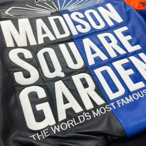 Puede incluir: Una chaqueta de cuero multicolor con paneles negros, azules y naranjas. La parte posterior muestra texto bordado en blanco que dice MADISON SQUARE GARDEN y THE WORLD'S MOST FAMOUS, con un logotipo de Avirex y l&iacute;neas gr&aacute;ficas sobre las letras.