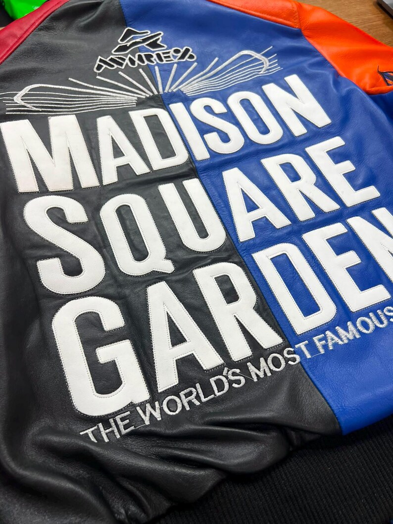 Puede incluir: Una chaqueta de cuero multicolor con paneles negros, azules y naranjas. La parte posterior muestra texto bordado en blanco que dice MADISON SQUARE GARDEN y THE WORLD'S MOST FAMOUS, con un logotipo de Avirex y l&iacute;neas gr&aacute;ficas sobre las letras.