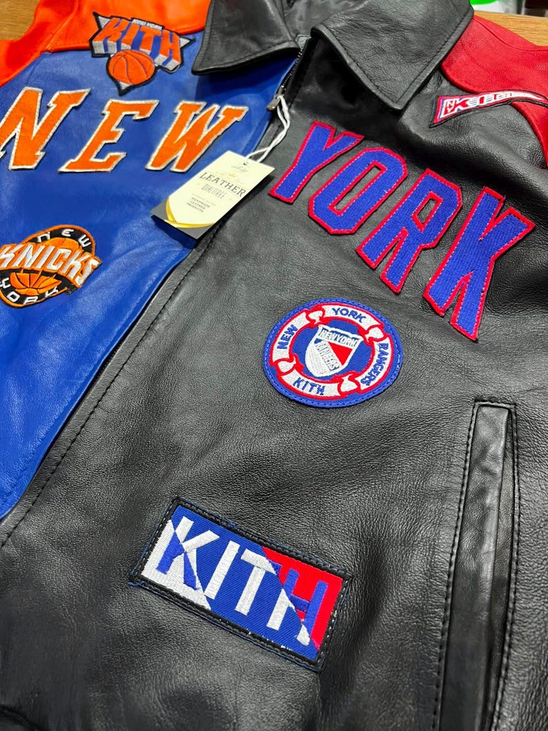 Puede incluir: Una chaqueta de cuero negra y azul con parches y letras bordadas en naranja, azul y blanco. La chaqueta muestra el texto NEW YORK, KITH y KNICKS, junto con un logotipo circular de los New York Rangers y una etiqueta de cuero adjunta.