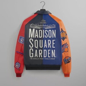 Puede incluir: Una chaqueta universitaria multicolor con un panel trasero azul oscuro con el texto blanco Madison Square Garden The World's Most Famous Arena. Las mangas son rojas y naranjas con varios parches y logotipos de equipos deportivos.