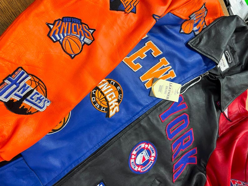 Puede incluir: Una colecci&oacute;n de chaquetas de cuero con paneles vibrantes en naranja, azul y negro. Las chaquetas est&aacute;n adornadas con logotipos bordados del equipo de baloncesto New York Knicks, letras grandes de New York y un parche circular de New York Rangers.