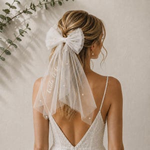 May include: A white tulle hair bow veil featuring scattered pearl embellishments and the embroidered text "bride to be" in cursive script. The sheer fabric is layered and gathered into a large bow shape, designed to be worn in the hair.
