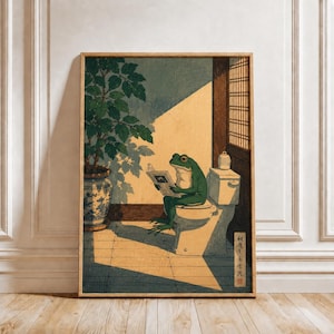 May include: A framed wall art print featuring a green frog sitting on a white toilet reading a book. To the left is a potted plant in a blue and white patterned ceramic vase. The scene is set in a room with a wooden lattice window and sunlight.