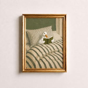 May include: A framed wall art print featuring a white duck with an orange beak sitting in a bed with striped bedding and pillows, holding a small green book open in its wings. The scene is set against a solid dark green wall background.
