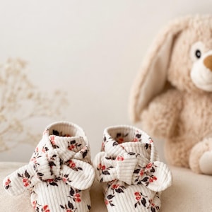 Puede incluir: Un par de patucos para bebé de pana color blanco roto con un estampado floral de pequeñas bayas rojas y hojas verde oscuro. Cada patuco tiene un detalle de lazo anudado decorativo en la parte delantera, punta redondeada y tejido acanalado.