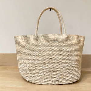 Può includere: Una borsa tote in paglia intrecciata color beige con un design ampio e aperto e due manici arrotondati abbinati. La borsa presenta un motivo a trama orizzontale consistente e strutturato su tutto il corpo, dall'aspetto naturale.