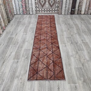 May include: A long, narrow runner rug featuring a deep terracotta red base colour with a repeating geometric pattern of dark brown lines forming interconnected triangles and diamonds across the entire surface of the rectangular textile floor covering.