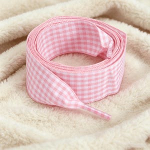 May include: A coiled shoelace featuring a light pink and white gingham check pattern. The shoelace is made of a woven fabric and includes a clear plastic aglet at the end, designed for threading through eyelets on footwear or other items.