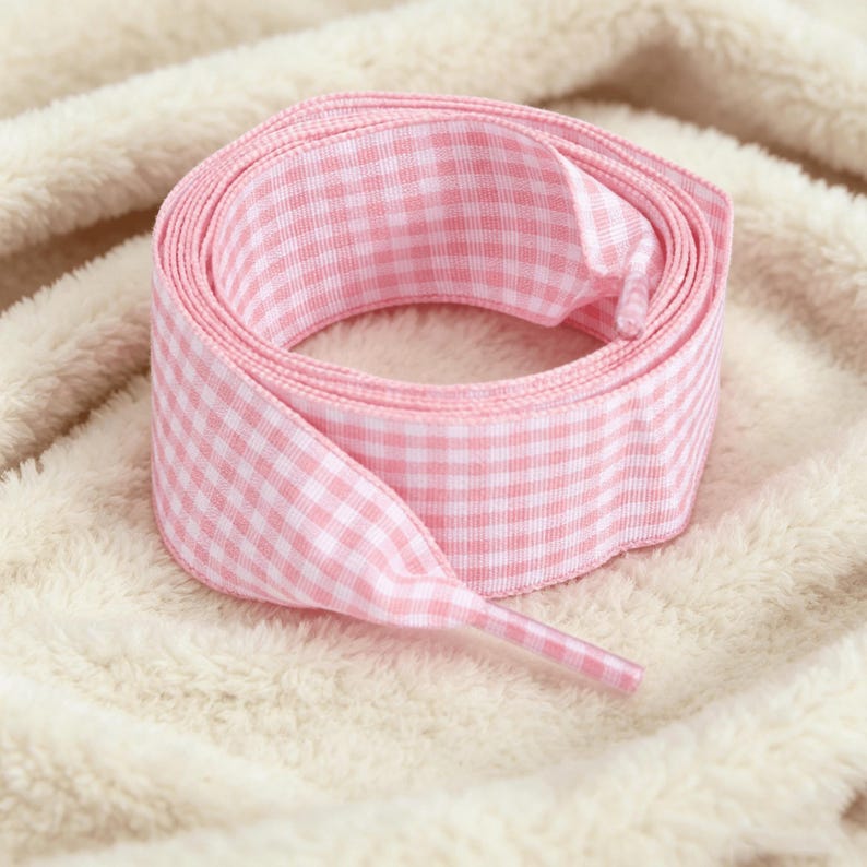 May include: A coiled shoelace featuring a light pink and white gingham check pattern. The shoelace is made of a woven fabric and includes a clear plastic aglet at the end, designed for threading through eyelets on footwear or other items.