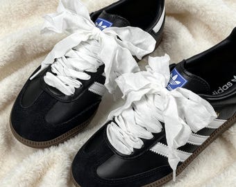 May include: A pair of black leather sneakers with white three-stripe branding, black suede toe overlays, and brown gum rubber soles. The shoes feature wide white fabric ribbon laces tied into large bows at the top, with blue and white tongue labels.