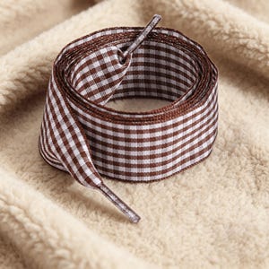 May include: A coiled shoelace featuring a brown and white gingham check pattern. The shoelace is made of a woven fabric and includes a plastic aglet at the end of the lace, which is designed for threading through eyelets on footwear.