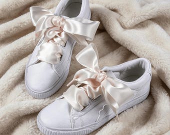 May include: A pair of white low-top sneakers featuring wide, pale pink satin ribbon laces tied into large, prominent bows. The shoes have a textured leather upper, a rounded toe, and a scalloped edge detail along the side panel near the laces.