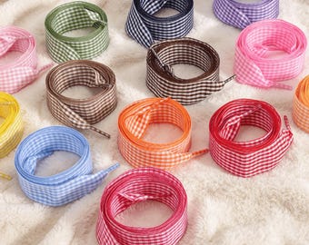 May include: A collection of flat shoelaces arranged in a circular pattern, each featuring a small-scale gingham check pattern. The set includes laces in various colors: green, navy blue, purple, pink, brown, orange, red, light blue, and yellow.