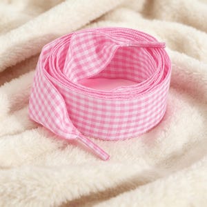 May include: A pair of flat shoelaces featuring a pink and white gingham checkered pattern. The laces are coiled into a circular shape, displaying the woven fabric texture and the plastic aglets at each end of the lace, designed for footwear.