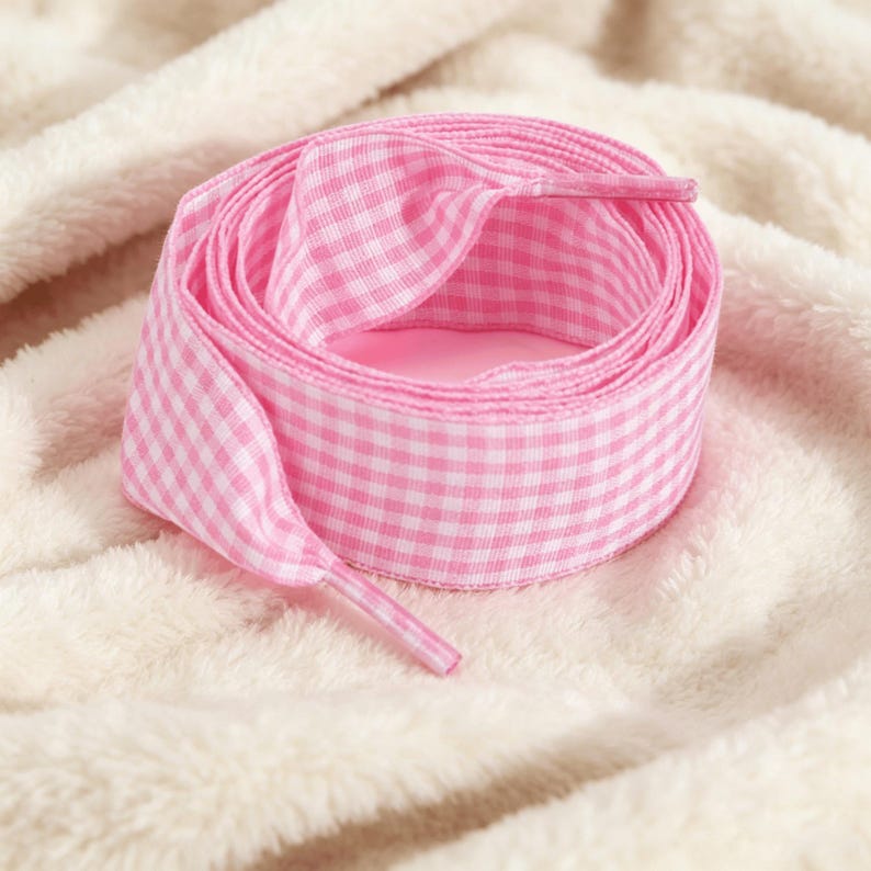 May include: A pair of flat shoelaces featuring a pink and white gingham checkered pattern. The laces are coiled into a circular shape, displaying the woven fabric texture and the plastic aglets at each end of the lace, designed for footwear.