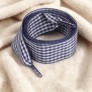 May include: A pair of flat shoelaces featuring a navy blue and white gingham check pattern. The laces are coiled into a circular shape and have matching navy blue plastic aglets at each end to prevent fraying and facilitate easy lacing.