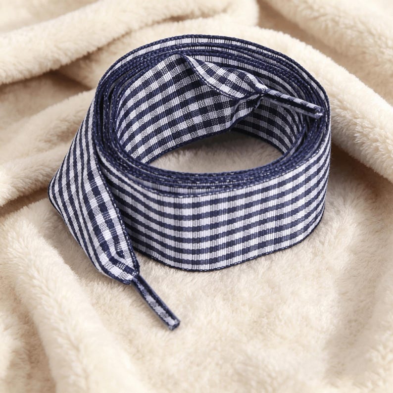 May include: A pair of flat shoelaces featuring a navy blue and white gingham check pattern. The laces are coiled into a circular shape and have matching navy blue plastic aglets at each end to prevent fraying and facilitate easy lacing.