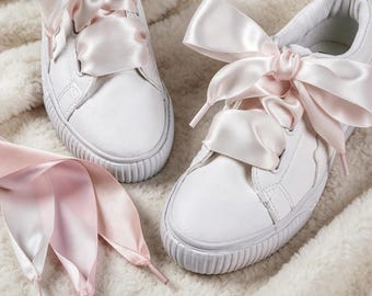 May include: A pair of white sneakers featuring wide, pale pink satin ribbon laces tied into large bows. The shoes have a textured white rubber sole and a smooth white upper. An extra set of pale pink satin ribbon laces is visible in the foreground.
