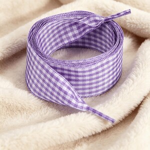 May include: A flat shoelace featuring a purple and white gingham check pattern. The shoelace is coiled into a circular shape, with both ends visible, showing the aglets at the tips of the fabric ribbon. The material has a woven texture.