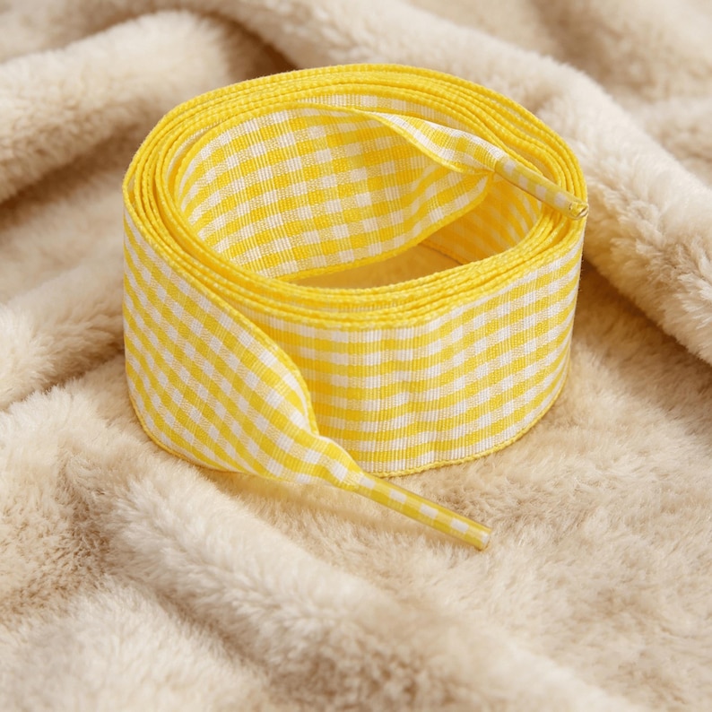 May include: A coiled shoelace featuring a bright yellow and white gingham check pattern. The shoelace is made of a flat, woven fabric and includes matching yellow and white checkered aglets at both ends to prevent fraying and aid in lacing.