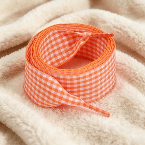 May include: A coiled length of flat shoelace featuring a vibrant orange and white gingham check pattern. The shoelace has a woven texture and includes a matching orange plastic aglet at the end, designed for securing through eyelets on footwear.