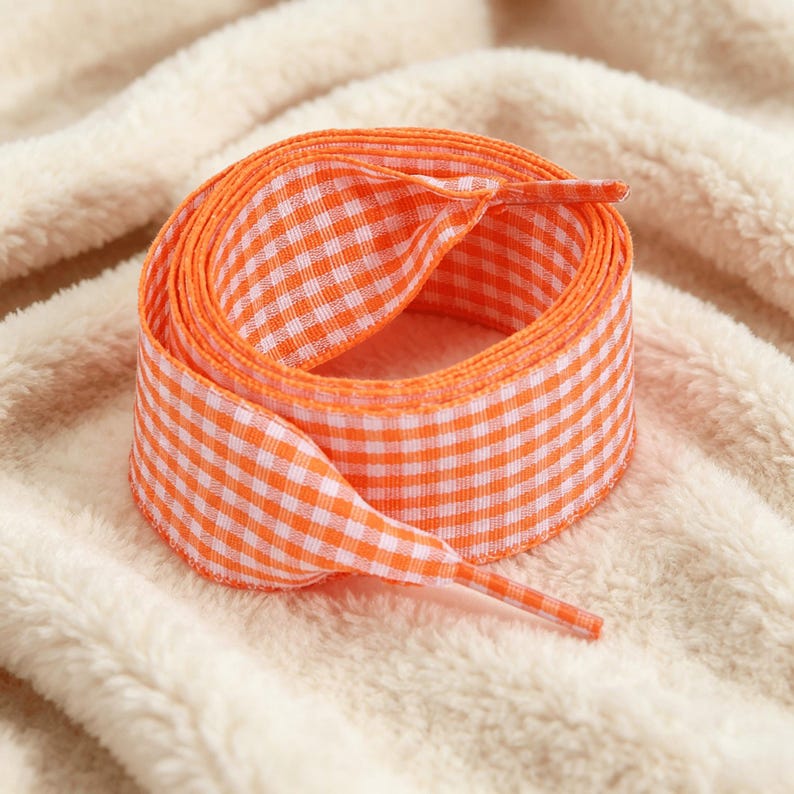 May include: A coiled length of flat shoelace featuring a vibrant orange and white gingham check pattern. The shoelace has a woven texture and includes a matching orange plastic aglet at the end, designed for securing through eyelets on footwear.