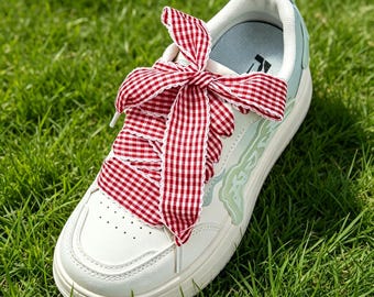 May include: A white sneaker featuring a thick, red and white gingham patterned ribbon lace tied in a large bow. The shoe has light blue accents along the side panels, a perforated toe box, and a white rubber sole with a pull tab at the heel.